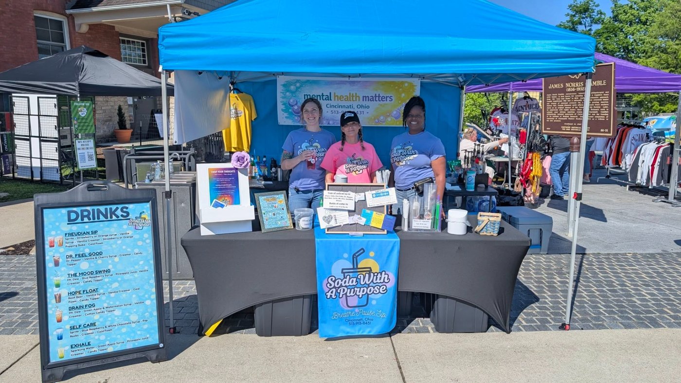 The three SWAP founders smiling behind their blue pop-up tent at a Cincinnati community event, with a Mental Health Matters banner and a DRINKS menu board